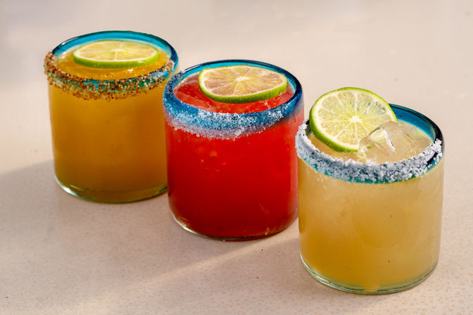 Vibrant trio of margarita-style cocktails in blue-rimmed short glasses with salted rims and lime slices — red and yellow drinks on a light surface.