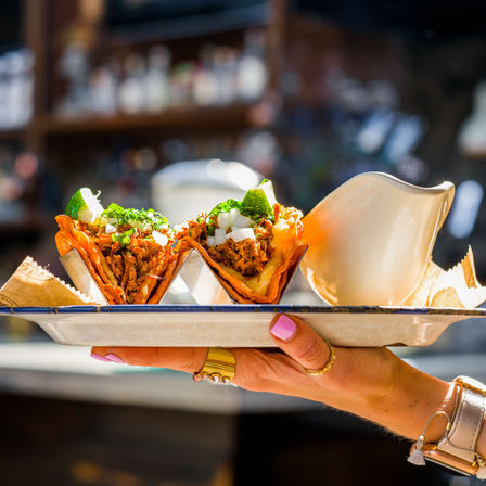 Two crispy shredded-meat tacos topped with cilantro, diced onion and lime wedges in a metal taco holder on an enamel tray, held over a blurred street-food/bar background