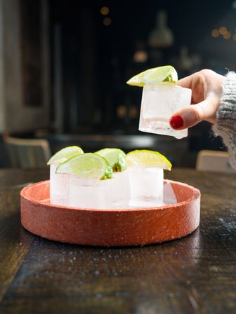 Hand with red nail polish lifting a lime-topped ice cube from a round tray of lime-garnished ice cubes on a wooden bar table