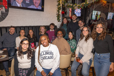 Cheerful group of diverse young adults posing at a festive indoor restaurant gathering with holiday garland; one person wears a 'University of Illinois Chicago' sweatshirt.