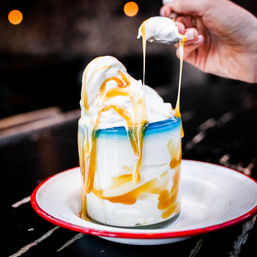 Drippy caramel cascading over creamy vanilla soft-serve in a glass jar, spoon lifting gooey caramel on a white enamel plate with red rim, bokeh lights in background