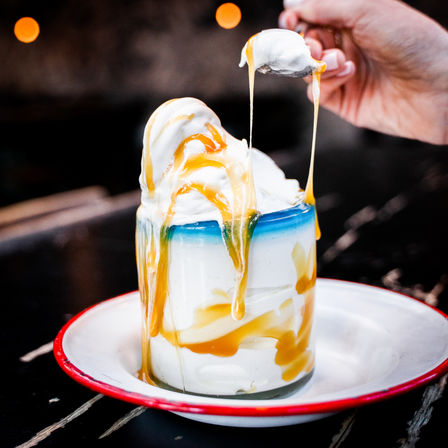 Drippy caramel cascading over creamy vanilla soft-serve in a glass jar, spoon lifting gooey caramel on a white enamel plate with red rim, bokeh lights in background