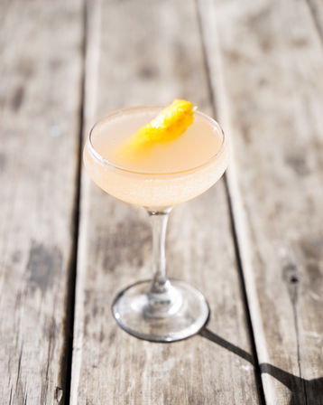 Sunlit pale-pink effervescent cocktail in a coupe glass topped with a bright peach peel, sitting on a weathered wooden table