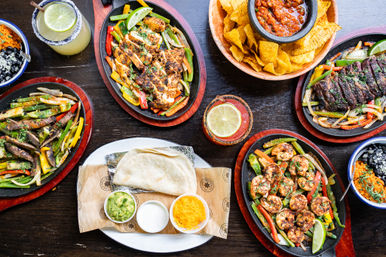 Top-down view of a colorful Tex-Mex spread: sizzling cast-iron fajitas with grilled chicken, steak and shrimp, warm tortillas with guacamole, sour cream and shredded cheese, tortilla chips and salsa, bowls of rice and beans, and margaritas on a dark wooden table.