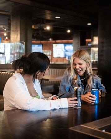 Two friends laughing at a modern urban bar booth, one in a denim jacket holding a blue can and the other in a white shirt with a tall can, casual nightlife scene with TVs and warm lighting in the background.