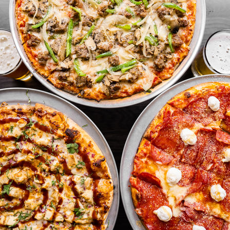 Overhead view of three large pizzas on metal trays on a dark wooden table: sausage, mushroom, onion and green pepper pizza; BBQ chicken pizza with red onion, cilantro and BBQ drizzle; pepperoni pizza topped with ricotta dollops, paired with two frothy beers.