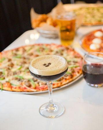 Espresso martini in a coupe glass topped with three coffee beans on a table with thin-crust pizzas, a pint of beer and a glass of red wine at a casual pizzeria.