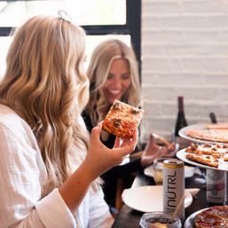 Two friends laughing at a cozy indoor pizzeria, one holding a square slice of cheesy tomato pizza over a table of pizza trays and drinks.