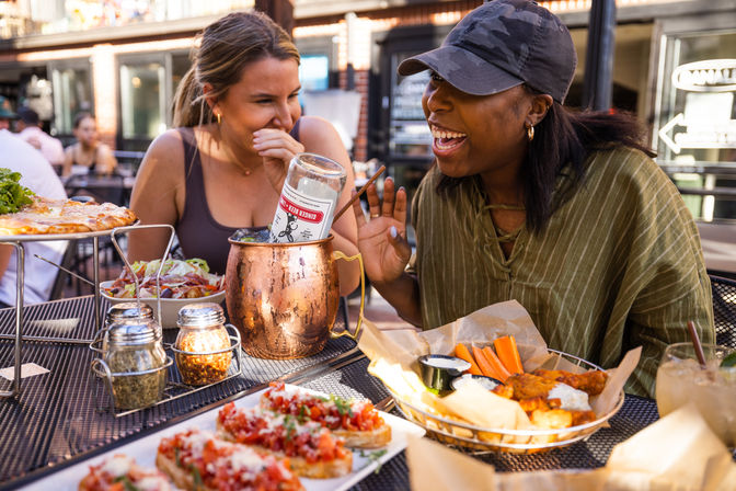 Two friends laughing over casual al fresco patio dining in the city, sharing bruschetta, pizza, salad, chicken wings with carrot sticks and a copper-mug cocktail on a sunny afternoon.
