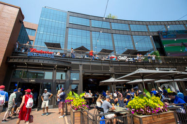 Sunny downtown sports bar with two-level outdoor patio and balcony, string lights and umbrellas, crowds of baseball fans eating, drinking and watching a game on a large exterior screen.