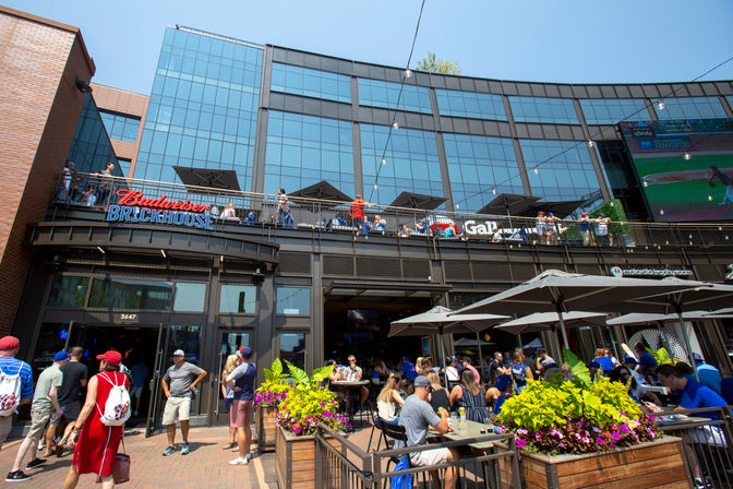 Sunny downtown sports bar with two-level outdoor patio and balcony, string lights and umbrellas, crowds of baseball fans eating, drinking and watching a game on a large exterior screen.
