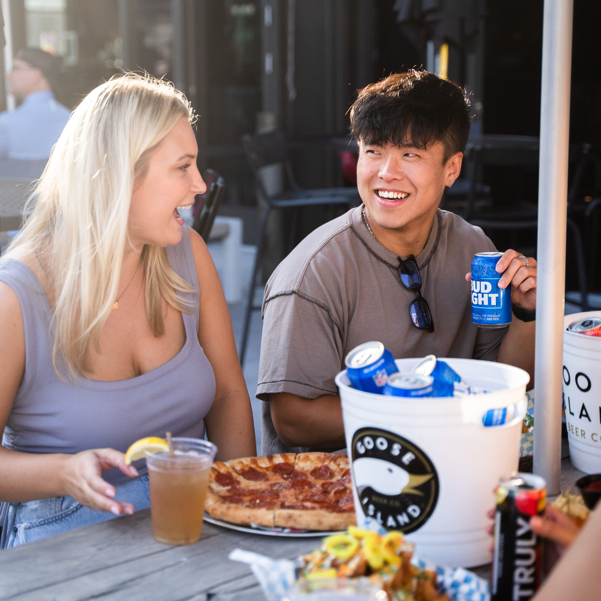 Two people laughing at an outdoor patio restaurant table, sharing a pepperoni pizza with a bucket of chilled beer cans and iced drinks during casual summer dining.