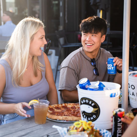 Two people laughing at an outdoor patio restaurant table, sharing a pepperoni pizza with a bucket of chilled beer cans and iced drinks during casual summer dining.