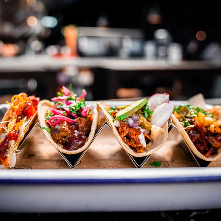 Close-up of assorted Mexican-style street tacos in a metal rack — soft corn tortillas filled with shredded and grilled meats, avocado, pickled onions, radish, cilantro and salsa, served on a restaurant counter.