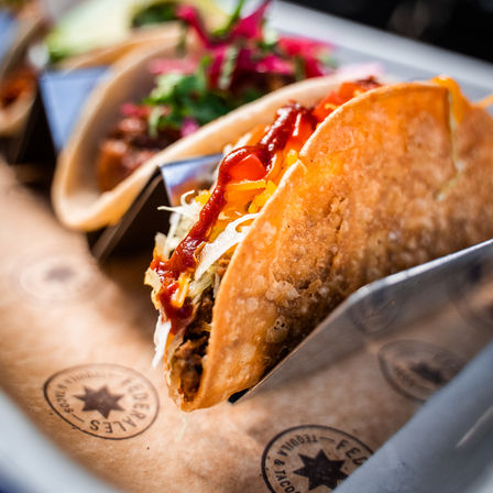 Close-up of a crispy hard-shell taco filled with seasoned meat, shredded lettuce and cheddar, drizzled with spicy red sauce — colorful street taco
