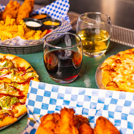 Close-up of a casual pub table spread: two pizzas, crispy chicken wings, crinkle fries with dipping sauces and two stemless glasses of red and white wine on blue checkered paper.