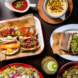 Overhead shot of a colorful Mexican taco platter with four street-style tacos, grilled corn queso, salsa, nachos and a lime-jalapeño margarita