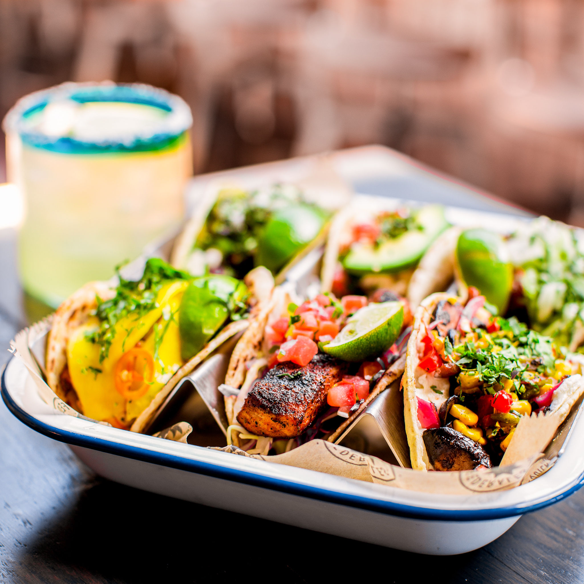 Assorted Mexican-style tacos in a tray — grilled seasoned fish, charred corn, pico de gallo, cilantro and lime wedges, with a salted-rim margarita blurred in the background.