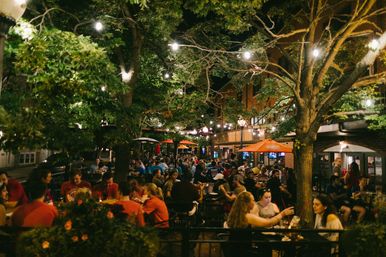 Lively urban outdoor dining patio at night with string lights and trees, crowded tables of people eating and drinking on a bustling streetscape