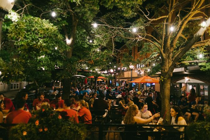 Lively urban outdoor dining patio at night with string lights and trees, crowded tables of people eating and drinking on a bustling streetscape