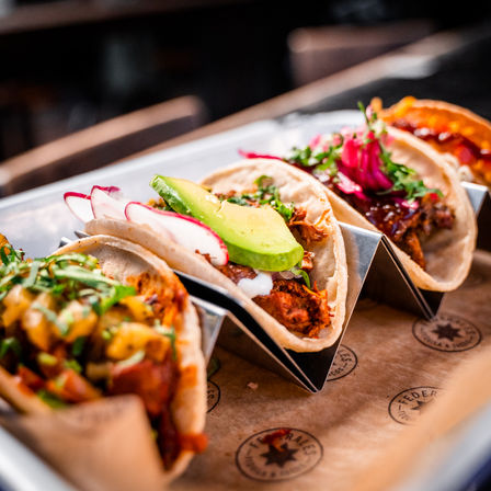 Three vibrant street tacos in a metal holder on a serving tray, filled with shredded meat, sliced avocado, pickled red onions, radish slices, cilantro and creamy sauce