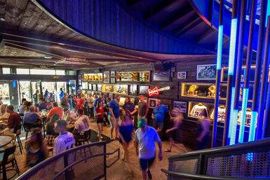 Crowded sports bar interior near a stadium entrance with fans in jerseys, glowing blue LED columns, neon sign, and display cases of baseball trophies and framed memorabilia.