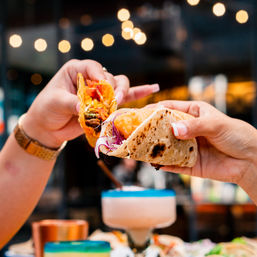 Two hands clinking tacos — a crunchy hard-shell taco with seasoned filling and a soft flour fish taco with purple cabbage — over a blue-rimmed cocktail on an outdoor patio with string lights.