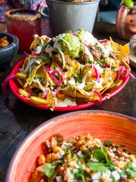 Loaded Mexican nachos on a red plate — tortilla chips piled with shredded meat, melted cheese, pickled red onions, jalapeños, dollops of guacamole and crema, served on a wooden table with drinks in the background.