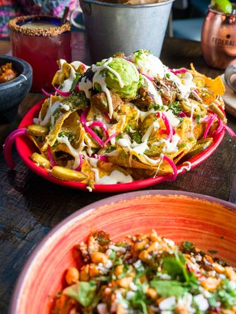 Loaded Mexican nachos on a red plate — tortilla chips piled with shredded meat, melted cheese, pickled red onions, jalapeños, dollops of guacamole and crema, served on a wooden table with drinks in the background.