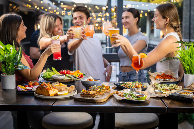 Group of friends toasting colorful cocktails over a wooden table of shareable summer dishes on a lit outdoor patio — al fresco dining with string lights.