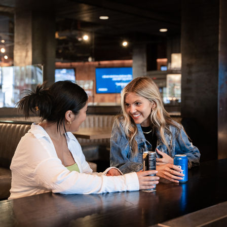 Two friends chatting and laughing over canned drinks at a cozy urban bar counter, one wearing a denim jacket and warm ambient lighting highlighting a modern wooden interior.