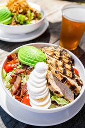 Grilled chicken Cobb salad bowl with sliced avocado, stacked hard-boiled egg, crispy bacon, cherry tomatoes and romaine on an outdoor wooden table with a glass of beer