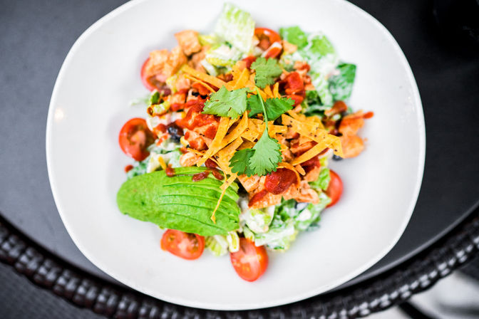Overhead view of a vibrant southwestern salad on a white plate with sliced avocado, cherry tomatoes, shredded tortilla strips, cilantro, grilled chicken and creamy dressing.