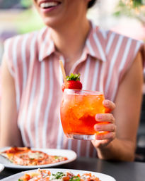 Bright orange strawberry spritz with ice and wooden stirrer held by a person in a pink-striped shirt above plates of pizza on an outdoor dining patio.