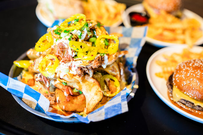 Smoky pulled-pork loaded fries topped with melted cheese, pickled banana peppers and green onions on blue checkered paper, with a sesame cheeseburger and crinkle-cut fries blurred in the background — casual American comfort food.