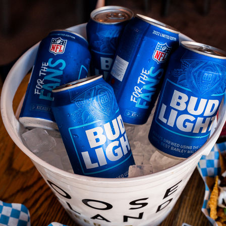 White bucket of ice-filled Bud Light cans with NFL "For the Fans" design on a wooden table, sports bar/tailgate vibe.