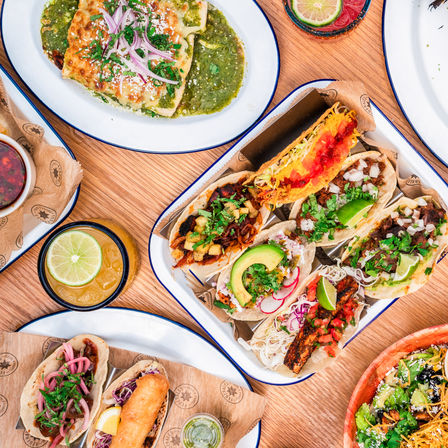 Overhead shot of a colorful Mexican spread: a taco platter with assorted street-style tacos (al pastor, fish, shredded beef, avocado), green enchilada, lime wedges, salsa and a fresh salad on a wooden table.