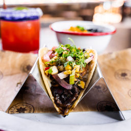 Close-up street-style taco in a metal stand with grilled meat, zesty charred corn salsa, pickled red onions and fresh cilantro, blurred margarita and side bowl in the background