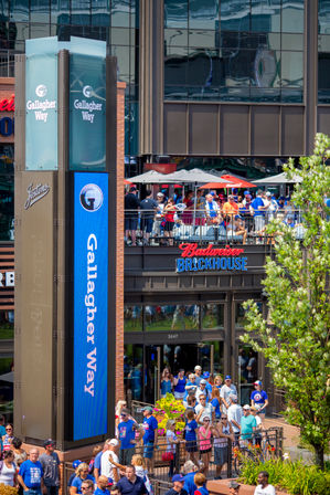 Busy summer stadium plaza with outdoor sports‑bar patio and balcony packed with fans in colorful team shirts under umbrellas, a tall digital column and reflective glass building in the background.