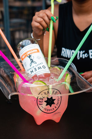 Pink tequila punch in a clear plastic party bucket with ice, lime slices, an inverted mini bottle and bright neon straws, hand with green nails reaching a straw