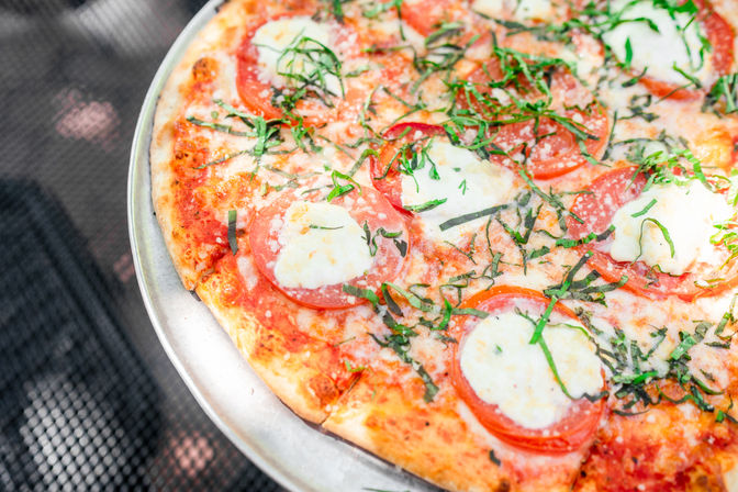 Sunlit thin-crust Margherita pizza on a metal stand with fresh mozzarella dollops, sliced tomatoes, shredded basil and grated Parmesan over bubbly tomato sauce