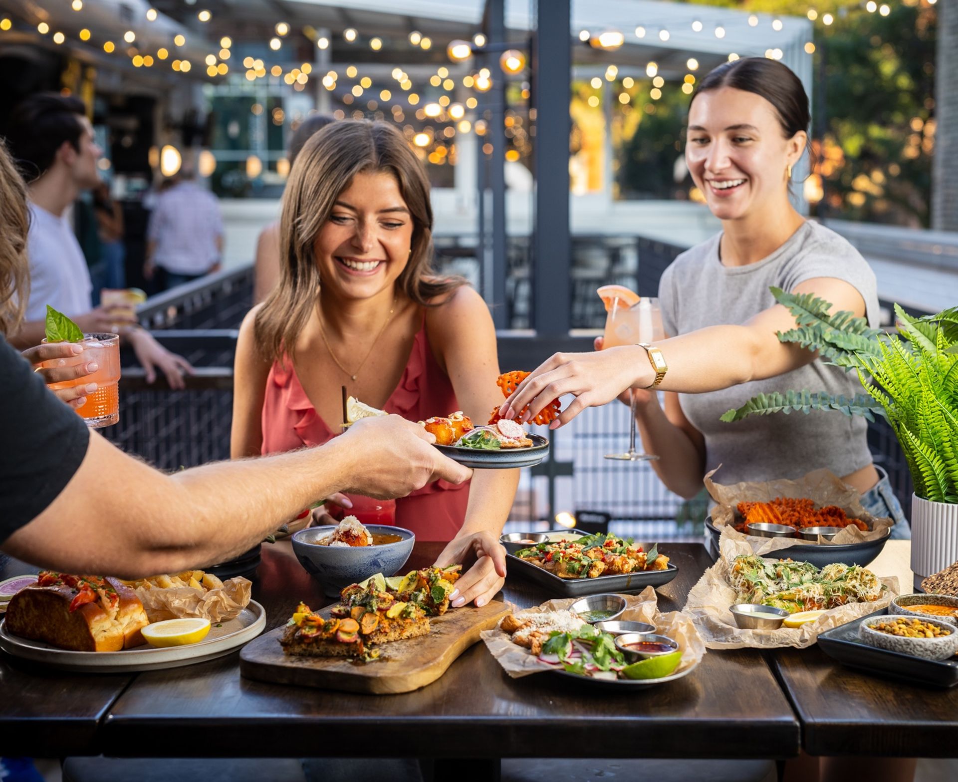 Smiling friends enjoying outdoor patio dining and happy hour—sharing cocktails and small plates like bruschetta, fries and salads on a wooden table under string lights.