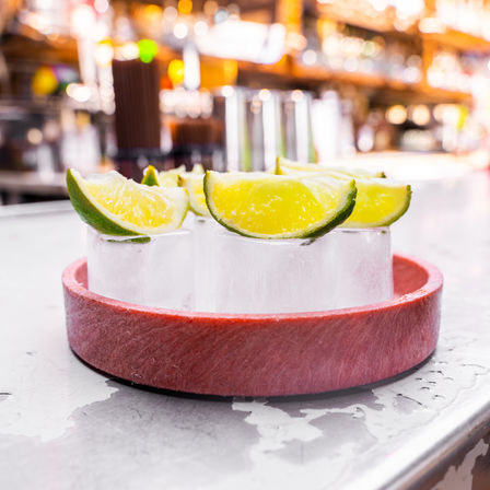 Close-up of bright lime wedges on cylindrical ice shot glasses arranged on a red tray on a bar counter, a refreshing cocktail garnish with warm bokeh of bottles and lights in the background
