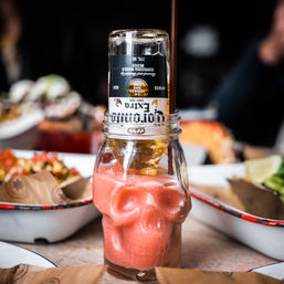 Skull-shaped glass with a pink frozen cocktail topped by an upside-down beer bottle and straw, surrounded by seafood plates and tortilla chips on a rustic table