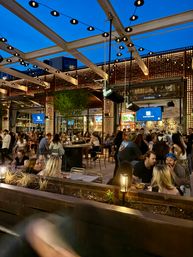 Rooftop bar patio at dusk — urban outdoor dining under a modern pergola with string lights, illuminated decorative wall panels, TVs, and crowds enjoying drinks and food.