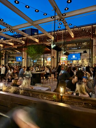 Rooftop bar patio at dusk — urban outdoor dining under a modern pergola with string lights, illuminated decorative wall panels, TVs, and crowds enjoying drinks and food.
