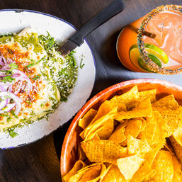 Top-down view of a Mexican plate with green salsa and melted cheese topped with pickled red onions and cilantro, a wooden bowl of tortilla chips, and a citrus cocktail with a chili-salt rim on a dark wood table.