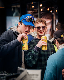 Two young men in casual jackets smiling and holding tall yellow cans at an outdoor downtown street festival under string lights
