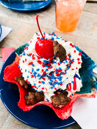 Patriotic red-white-blue ice cream sundae in a colorful waffle bowl topped with whipped cream, red and blue star sprinkles, chocolate teddy cookies and a maraschino cherry