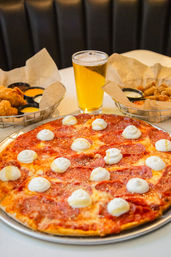 Crispy thin-crust pepperoni pizza dotted with ricotta dollops on a metal tray, served with a pint of beer and baskets of fried appetizers and dipping sauces on a casual pizzeria table.